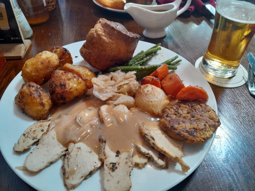A Sunday roast with chicken, roasties, Yorkshire pudding, a small selection of vegetables and a sausage patty.